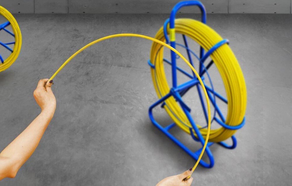 Pilot do przeciągania kabli EXTRALINK EX.16026 Dwie ręce zgięte w łuk trzymają żółty przewód, a w tle widać niebieski stojak z nawiniętą resztą przewodu na szarym podłożu