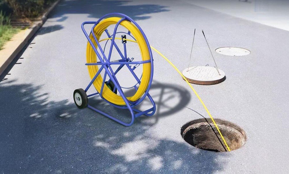 Pilot do przeciągania kabli EXTRALINK EX.13919 Niebieski stojak z żółtym przewodem ustawiony na asfalcie obok otwartego włazu kanalizacyjnego, do którego wprowadzony jest przewód