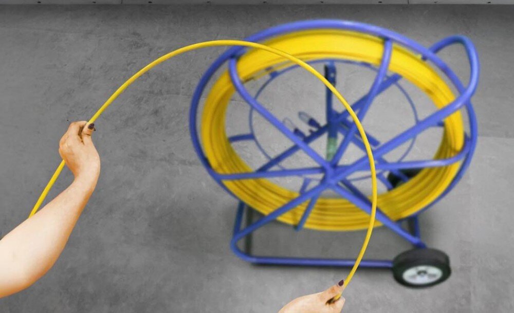 Pilot do przeciągania kabli EXTRALINK EX.13919 Dwie ręce zgięte w łuk trzymają żółty przewód, a w tle widać niebieski stojak z nawiniętą resztą przewodu na szarym podłożu