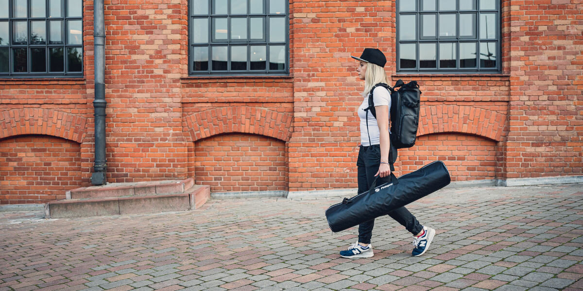 Statyw FOTOPRO DV-2, fotopro, niezbędne, akcesoria, pokrowiec, transport, kobieta idzie przez ulicę trzymając spakowany statyw w pokrowcu