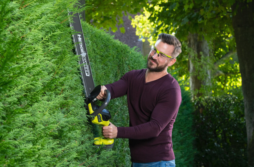 Nożyce do żywopłotu RYOBI RY18HT55A-120 550mm 18V akumulatorowe ogród osoba przycina żywopłot w tle drzewa krzewy wygoda pielęgnacja ogrodu swoboda ruchu
