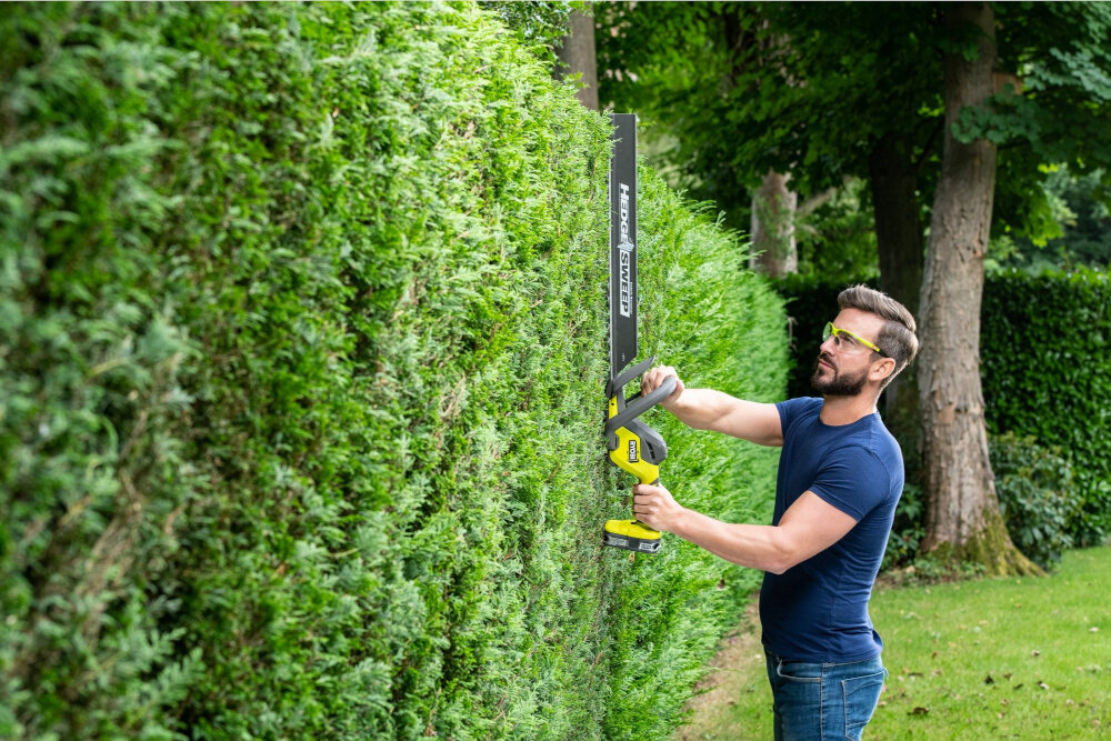 Nożyce do żywopłotu RYOBI RY18HT55A-120 550mm 18V akumulatorowe ogród osoba stoi przycina żywopłot w tle drzewa krzewy silnik szczotkowy stabilna i przewidywalna praca wspiera równomierne przenoszenie mocy na ostrze płynność ruchów ogranicza nagłe spadki wydajności