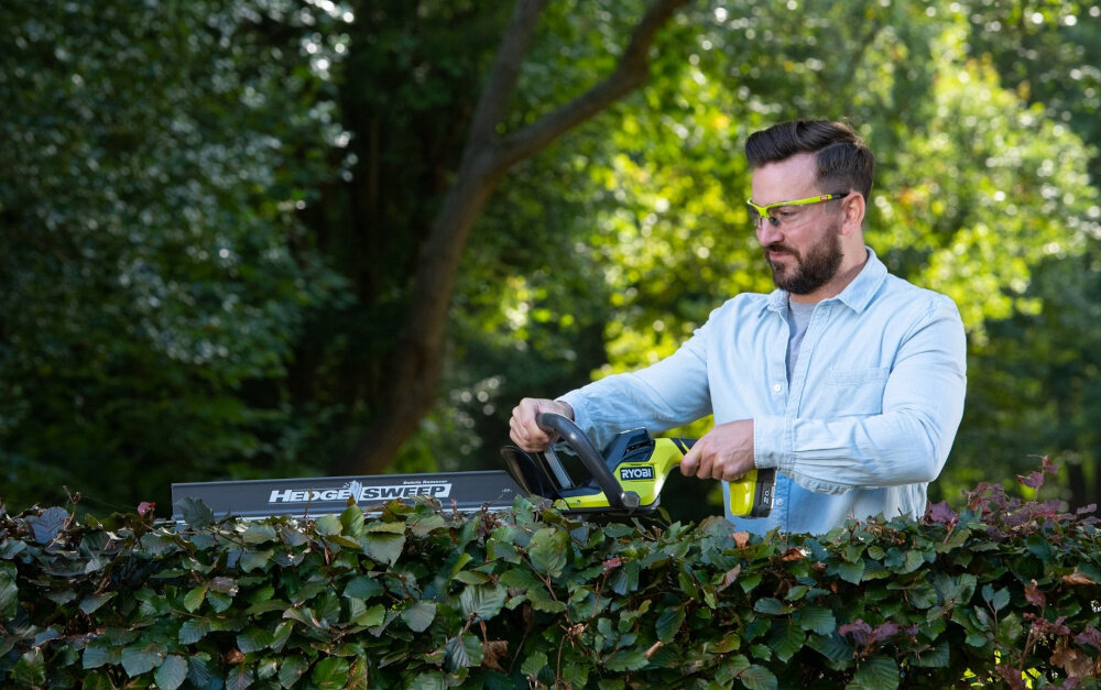 Nożyce do żywopłotu RYOBI RY18HT55A-120 550mm 18V akumulatorowe ogród osoba przycina żywopłot w tle drzewa krzewy długość ostrza 55 cm maksymalna zdolność cięcia do 24 mm większa efektywność działań równomierne przycięcie na całej długości roślin