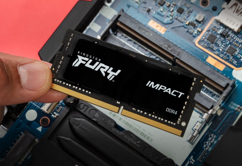Pamięć RAM KINGSTON Fury Impact 32GB 3200MHz Dłoń trzymająca pojedynczy moduł pamięci z logo 'FURY' i napisem 'IMPACT DDR4' nad otwartym wnętrzem laptopa, napięcie, działanie