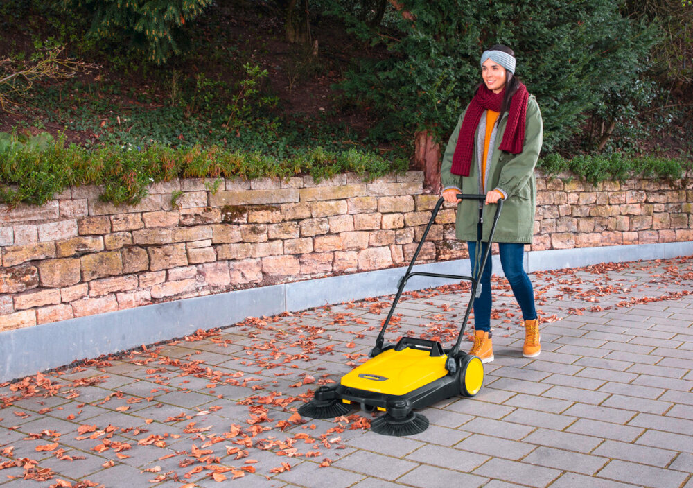 Zamiatarka ręczna KARCHER S4 Twin 2-in-1 1.766-365.0 podjazd chodnik osoba sprząta liście z ziemi w tle drzewa krzewy szerokość robocza do 680 mm mocna szczotka walcowa wydajność powierzchniowa do 2400 m² na godzinę znacznie mniej przejść potrzebnych do oczyszczenia dużych powierzchni