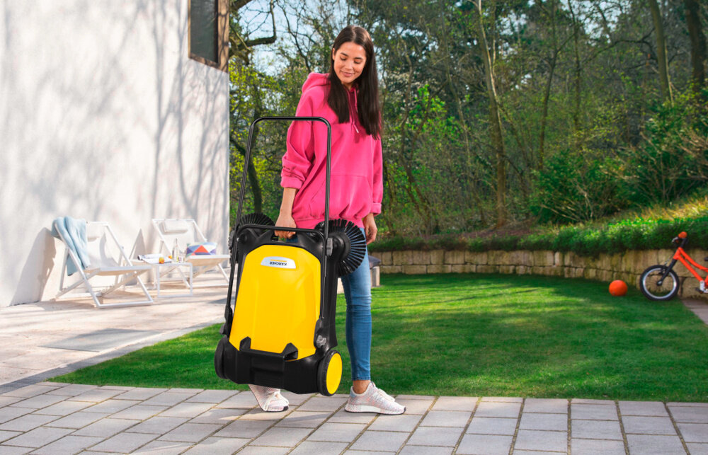 Zamiatarka ręczna KARCHER S4 Twin 2-in-1 1.766-365.0 ogród osoba niesie zamiatarkę w dłoni w tle trawnik dom krzewy drzewa waga około 10,2 kg obudowa z tworzywa sztucznego poprawia manewrowanie i zmniejsza zmęczenie użytkownika,