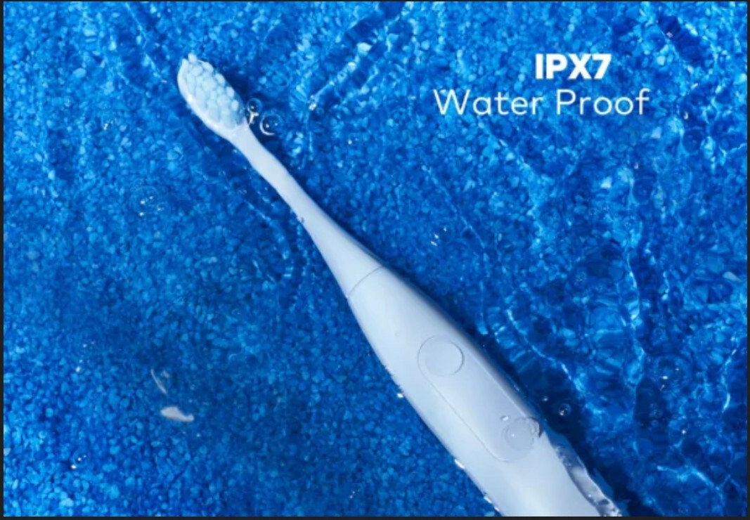 Na obrazie widoczna jest biała szczoteczka soniczna zanurzona w wodzie. Obok niej znajduje się tekst: \'IPX7 Water Proof\'.