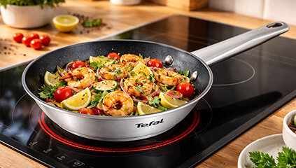 Na obrazie widoczna jest patelnia z potrawą składającą się z krewetek, plasterków cytryny i pomidorków koktajlowych, umieszczona na płycie indukcyjnej. Na patelni znajduje się logo \'Tefal\'.