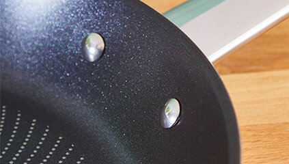 Zbliżenie na wnętrze czarnej patelni z metalowymi nitami i uchwytem. Powierzchnia patelni ma drobne, jasne kropki.