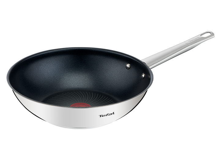 Na obrazie widoczna jest patelnia wok z ciemnym wnętrzem i jasnym uchwytem, z logo \'Tefal\' na boku. W środku patelni znajduje się czerwony okrąg z wzorem w kropki.