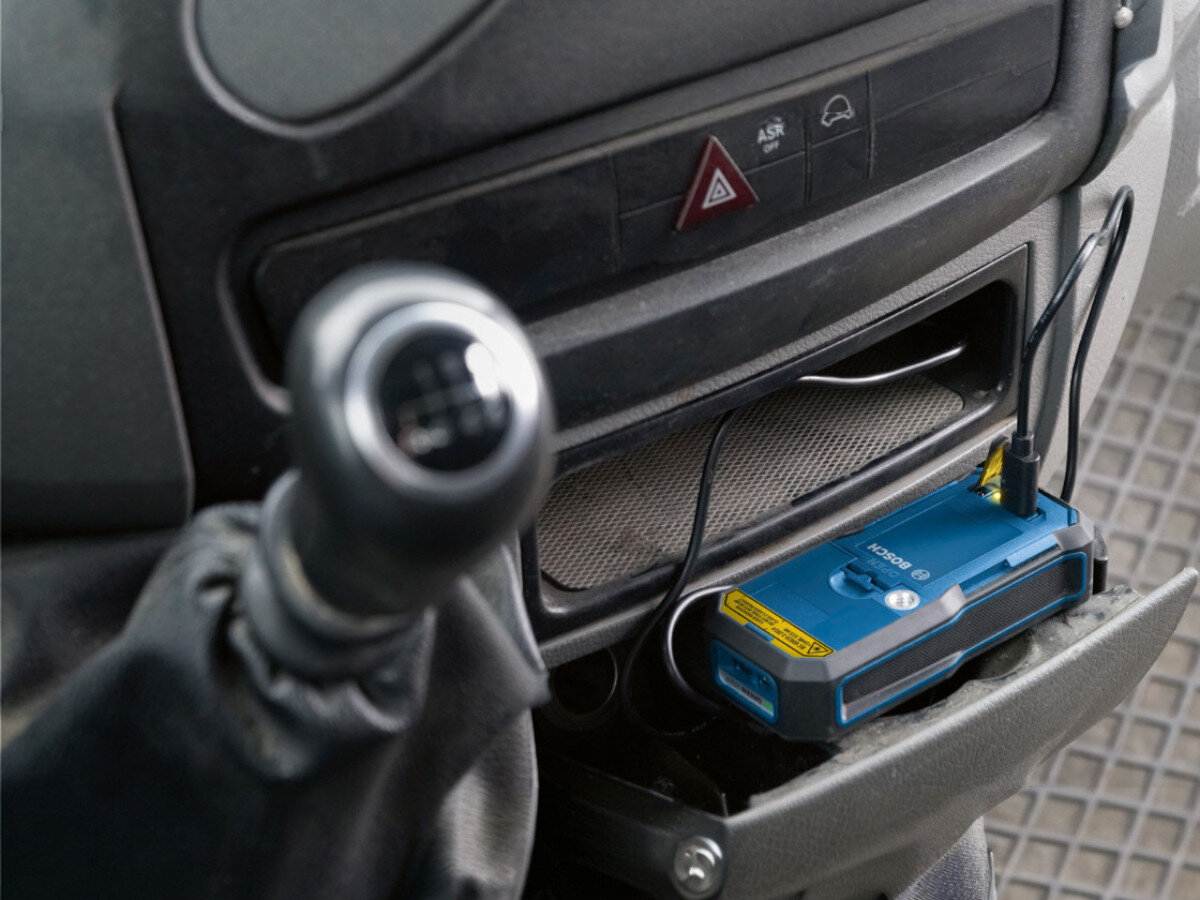 Wnętrze samochodu z widocznym niebieskim urządzeniem marki BOSCH podłączonym do gniazda ładowania. Obok znajduje się dźwignia zmiany biegów.