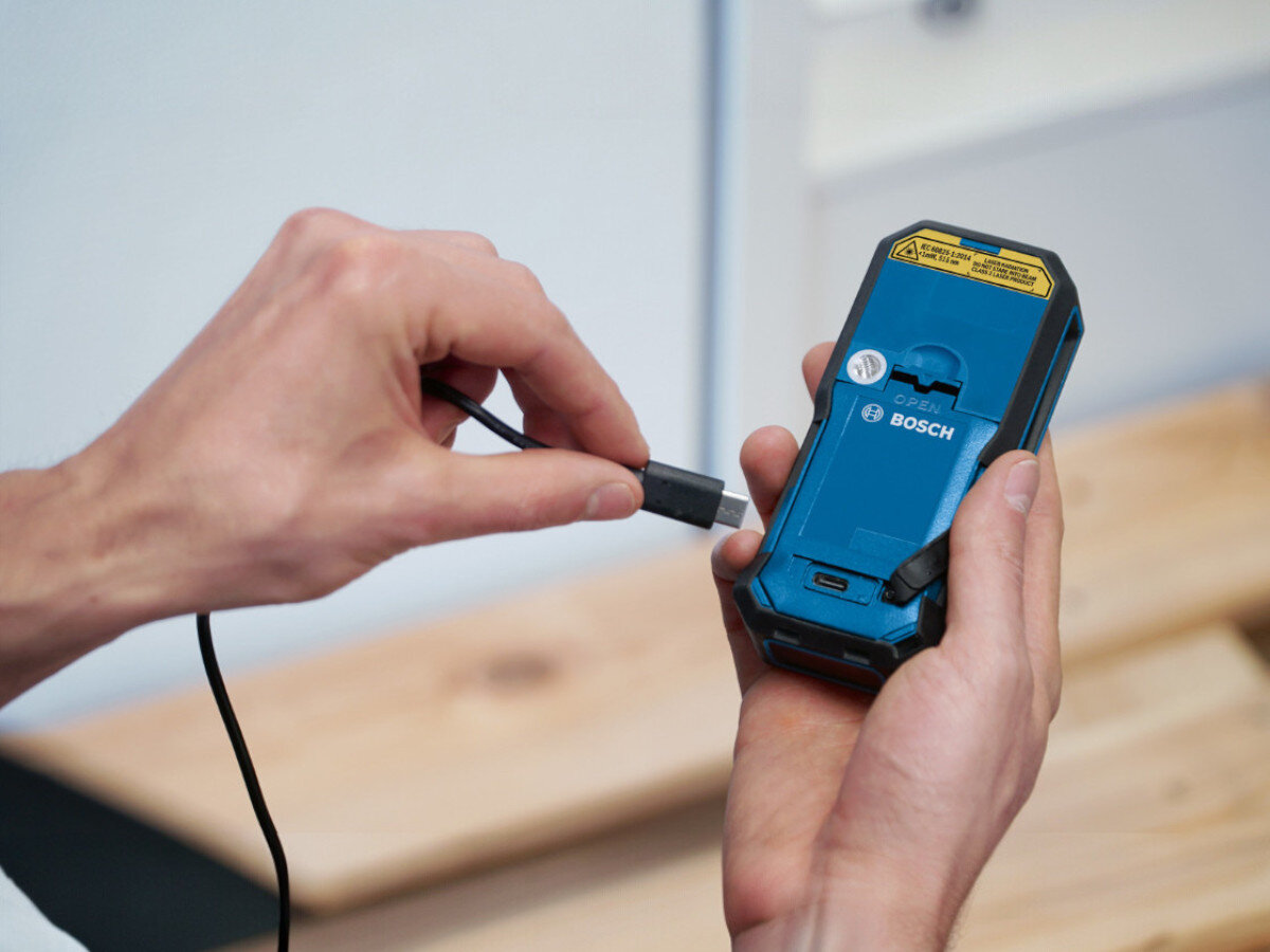 Osoba trzyma niebieskie urządzenie marki BOSCH i podłącza do niego kabel. Na urządzeniu widoczna jest naklejka z ostrzeżeniem oraz logo BOSCH.