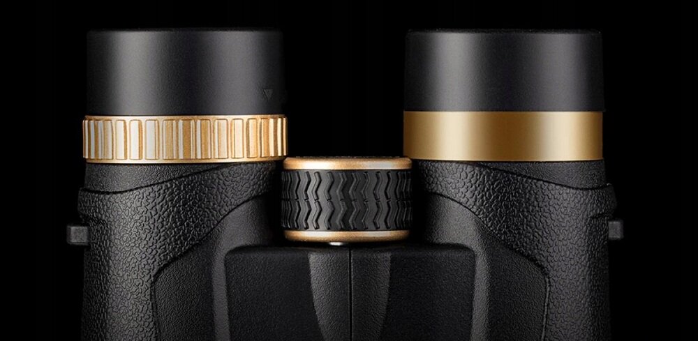 Zbliżenie na górną część lornetki z czarnymi i złotymi elementami. Centralne pokrętło ma teksturowaną powierzchnię.