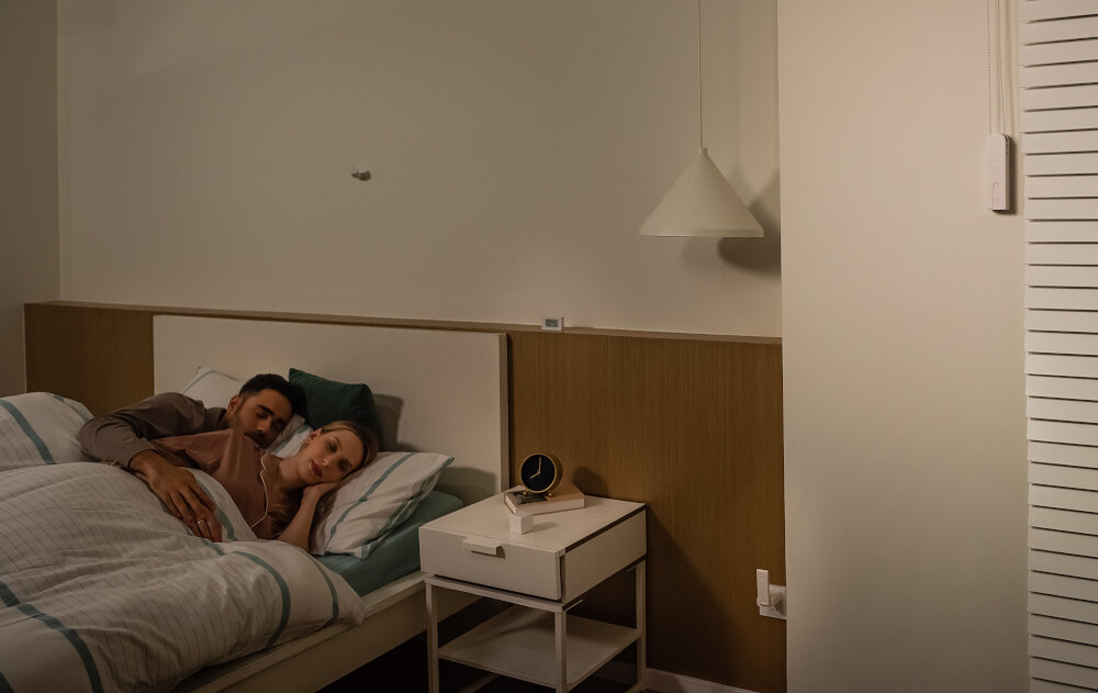 Sypialnia z łóżkiem, na którym leżą dwie osoby. Obok łóżka znajduje się stolik nocny z zegarem i lampką, a na ścianie wisi lampa sufitowa.