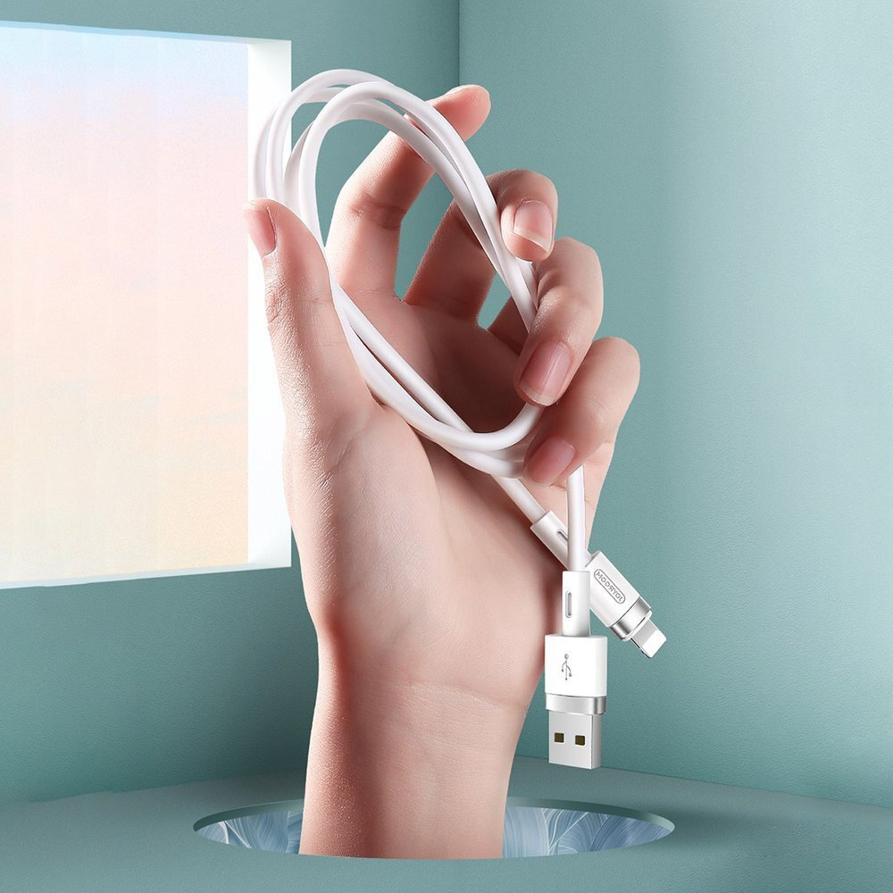 Osoba trzyma biały kabel USB z końcówką Lightning, widoczny na tle jasnoniebieskiej ściany z oknem. Na kablu znajduje się logo producenta.