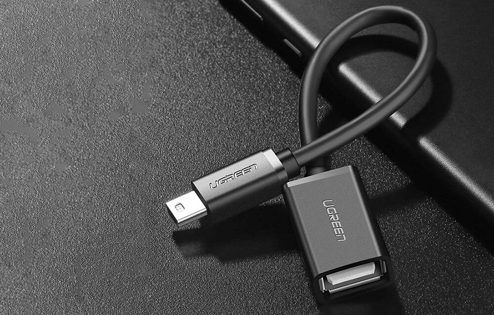 Czarny adapter OTG z logo \'UGREEN\' na obu końcach, z jednej strony zakończony wtykiem USB, a z drugiej gniazdem USB, leży na ciemnej powierzchni.