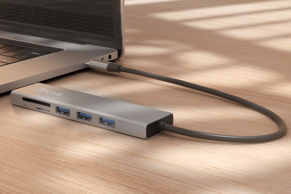 Hub TRUST Halyx 24191 Szary hub podłączony przewodem do laptopa leżący na drewnianym blacie w ostrym świetle dziennym