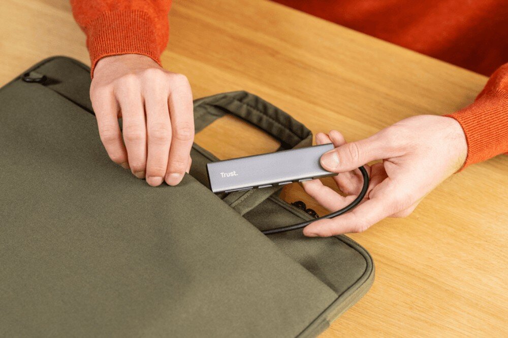 Hub TRUST Halyx 24191 Dłonie w pomarańczowym swetrze chowające szary hub do bocznej kieszeni zielonej torby na laptopa