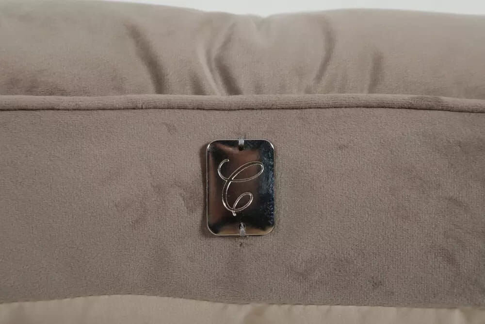 Legowisko dla kota ZOLUX Chambord z pikowaniem chesterfield i metalową aplikacją C Zbliżenie na detal poduszki: metalowa, prostokątna blaszka z grawerowaną literą 'C' (prawdopodobnie logo kolekcji) przymocowana do bocznej, welurowej ściany legowiska.