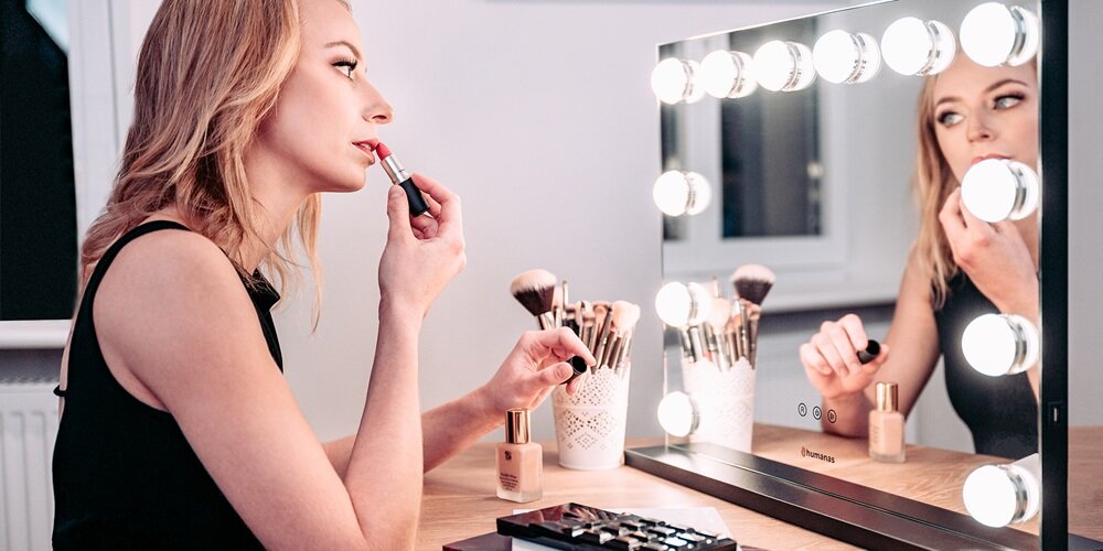 Kobieta nakłada szminkę przy prostokątnym lustrze Humanas z lampkami; na blacie stoją pędzle i kosmetyki