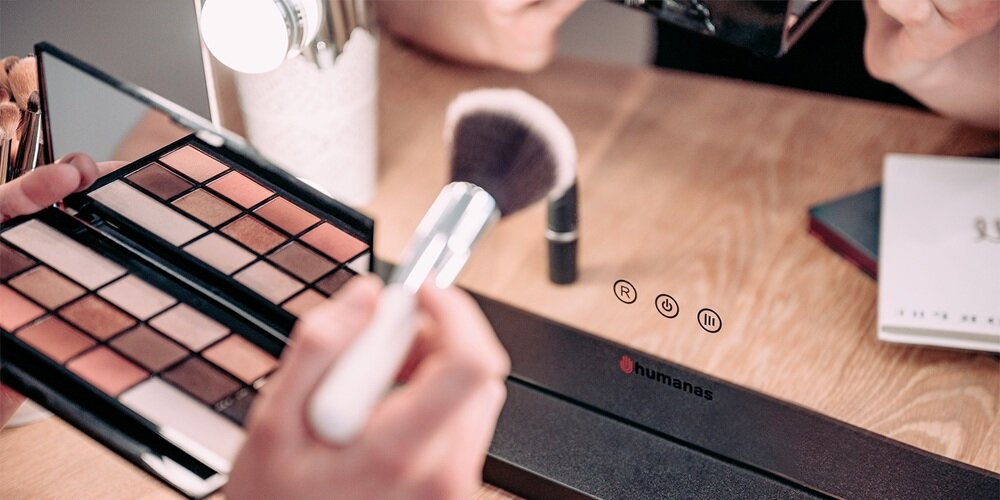 Zbliżenie na podstawę lustra Humanas z ikonami dotykowymi; obok paleta cieni i pędzel do makijażu na blacie