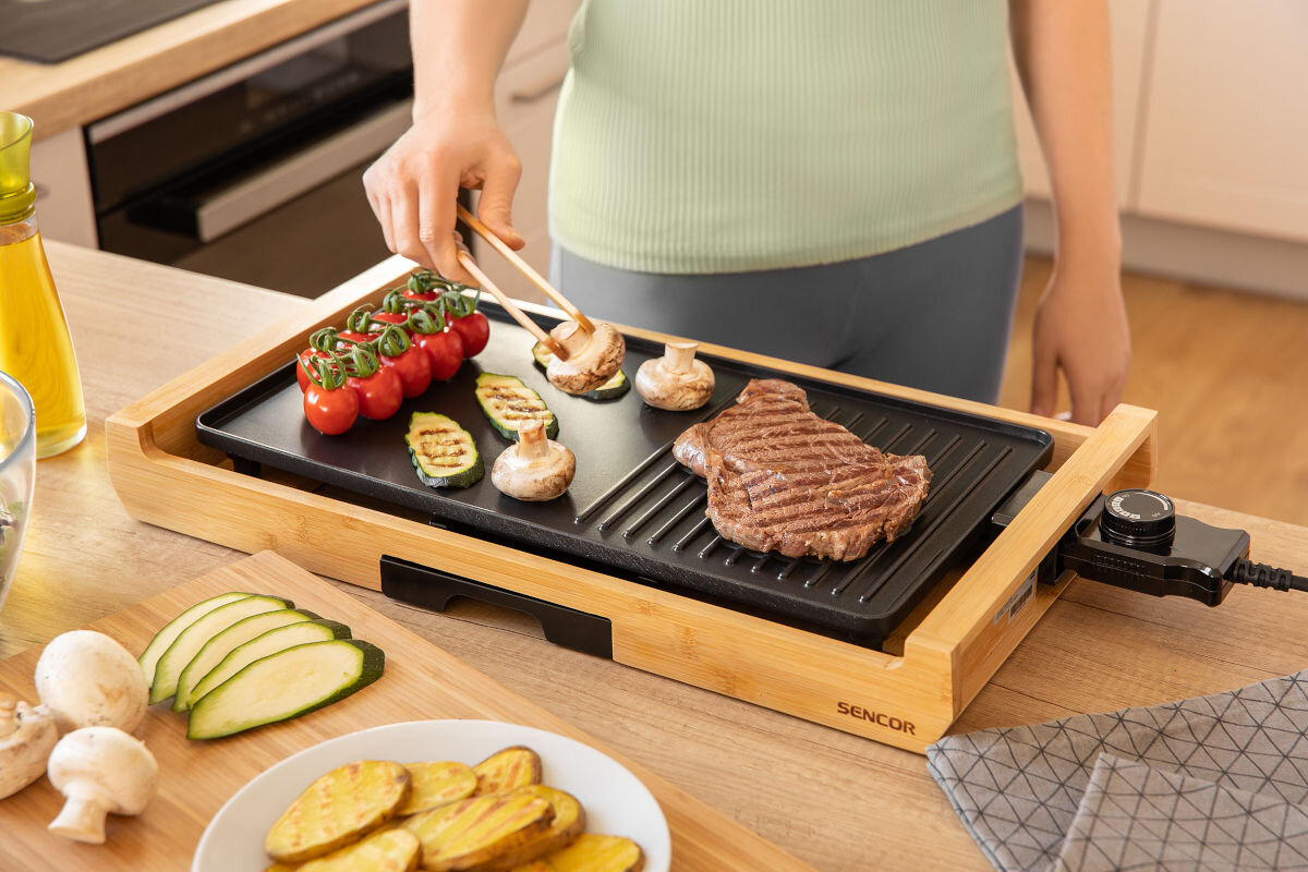 Grill elektryczny SENCOR SBG 206BK, kuchnia, gotowanie, grillowanie, dom, grill stoi na blacie w kuchni, w tle osoba obraca potrawy smażące się na grillu