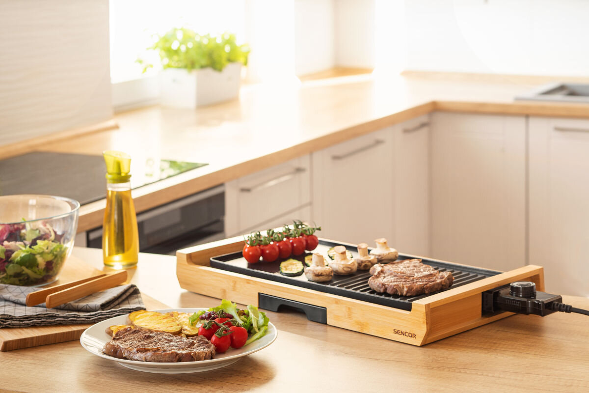 Grill elektryczny SENCOR SBG 206BK, zaprojektowane, antypoślizgowe, nóżki, nieprzywierające, grill stoi na blacie w kuchni, w tle talerze z przygotowanymi potrawami