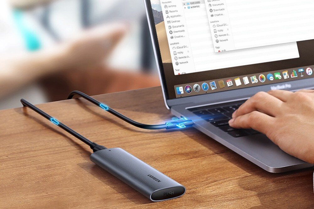 Obudowa dysku UGREEN CM400 Perspektywa z boku na szary dysk zewnętrzny podłączony do laptopa, na którego ekranie wyświetlone jest okno menedżera plików, a na kablu naniesiono graficzne strzałki i ikony sugerujące przesył danych