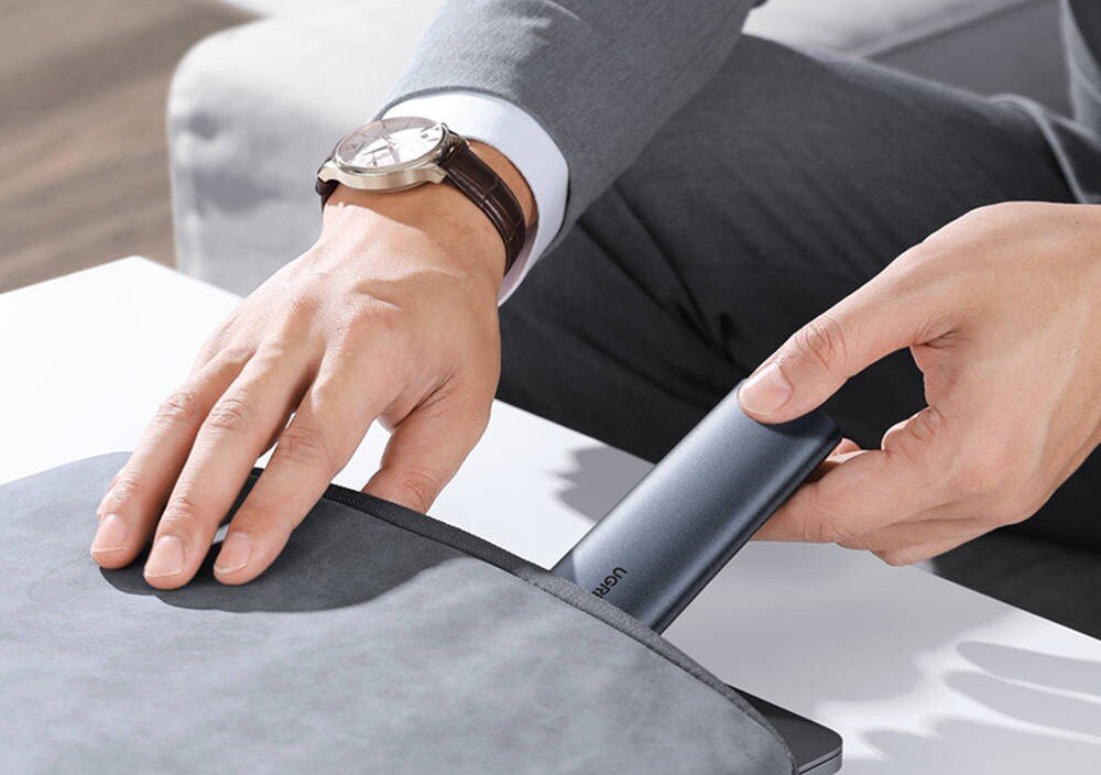 Obudowa dysku UGREEN CM400 Mężczyzna w szarym garniturze z zegarkiem na ręku wsuwa płaski, szary dysk zewnętrzny do bocznej kieszeni szarego etui na laptopa