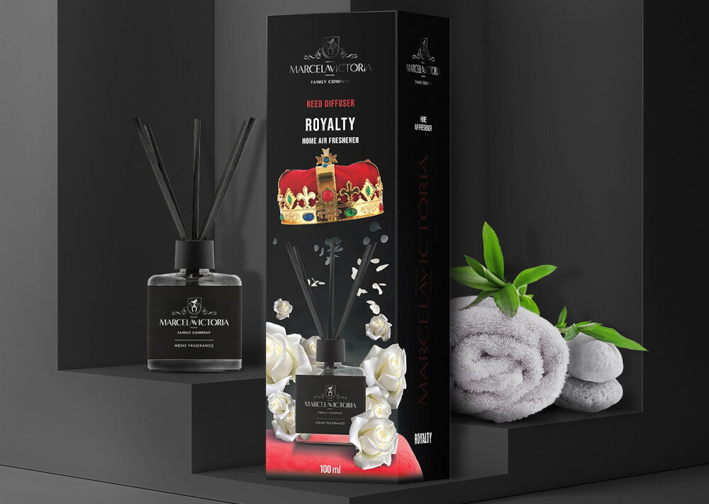 Na obrazie widoczny jest produkt \'Patyczki zapachowe MARCELA VICTORIA Royalty 100 ml\' w eleganckim czarnym opakowaniu z białymi różami i koroną. Obok opakowania znajduje się flakon z patyczkami, a w tle widoczne są ręczniki i zielone liście.