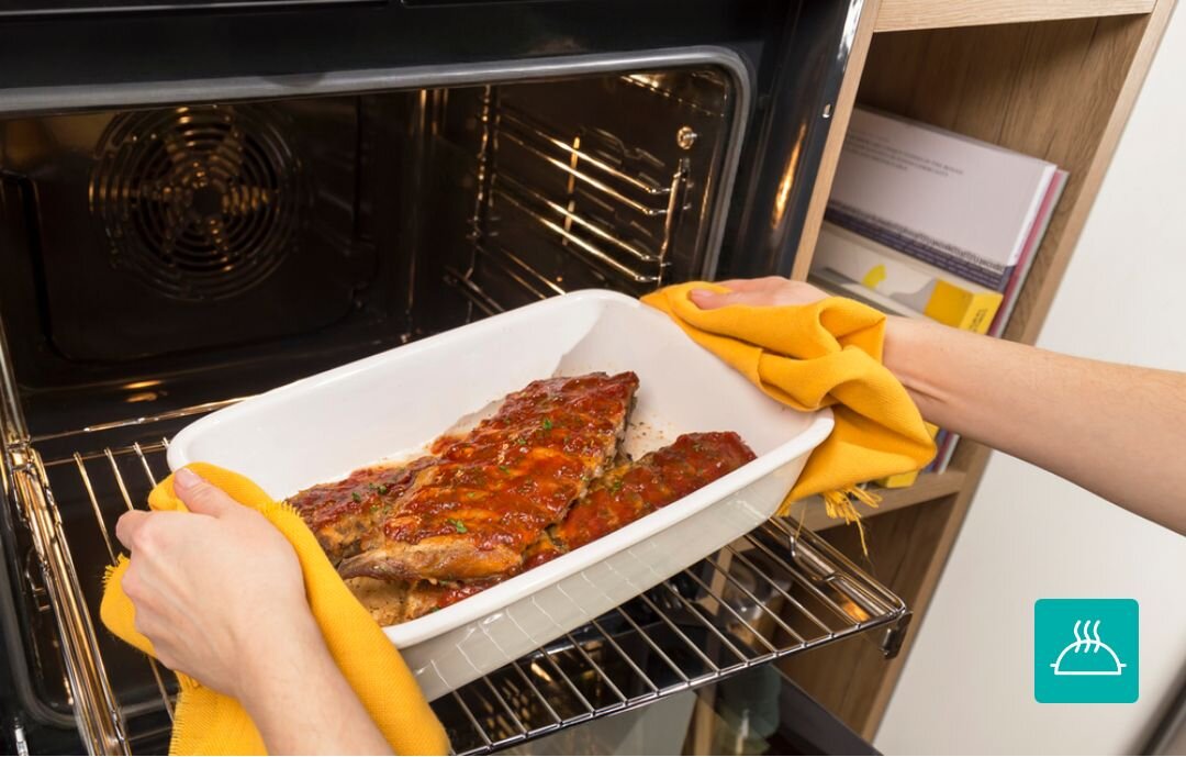 PIEKARNIK GORENJE BO6735E05B Wyjmowanie gorącej potrawy w naczyniu żaroodpornym z piekarnika przy użyciu kuchennych rękawic. Zdjęcie akcentuje bezpieczeństwo i wygodę użytkowania