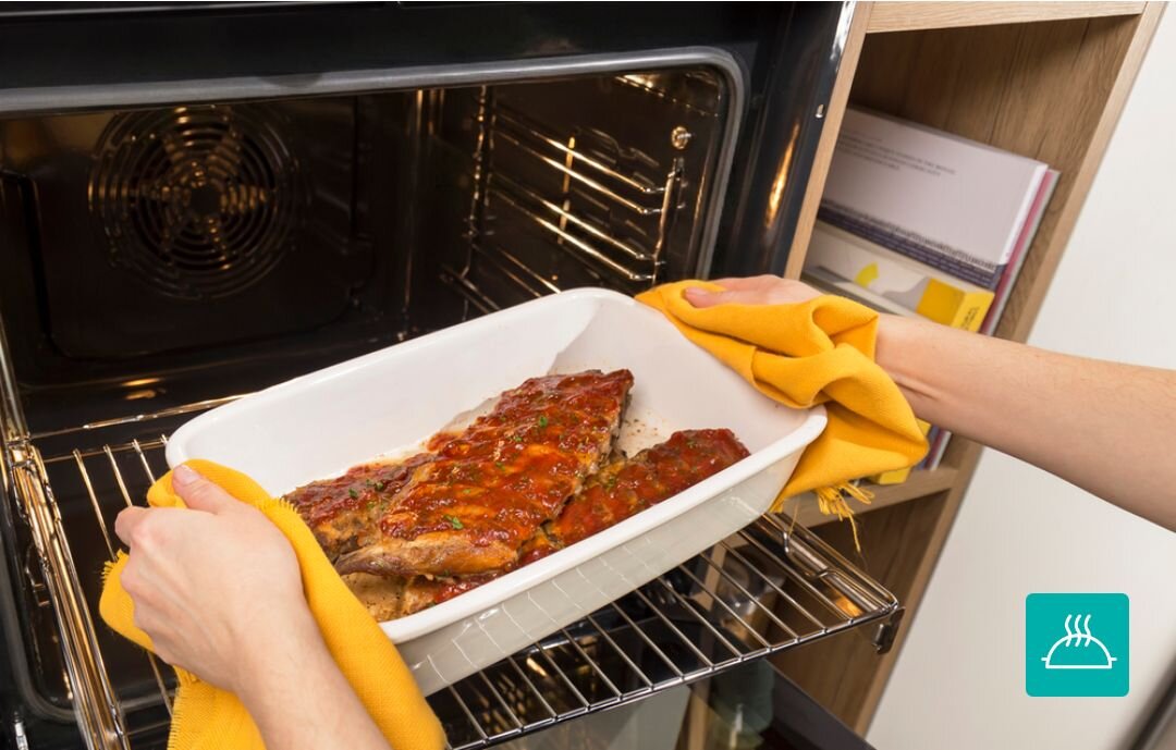 PIEKARNIK GORENJE BSA6737E15BG Wyjmowanie gorącej potrawy w naczyniu żaroodpornym z piekarnika przy użyciu kuchennych rękawic. Zdjęcie akcentuje bezpieczeństwo i wygodę użytkowania