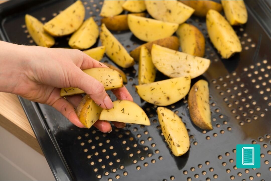 PIEKARNIK GORENJE BSA6737E15BG Ręka układa przyprawione ziemniaki na perforowanej blasze do pieczenia. Zdjęcie pokazuje przygotowanie potrawy z myślą o równomiernym dopieczeniu