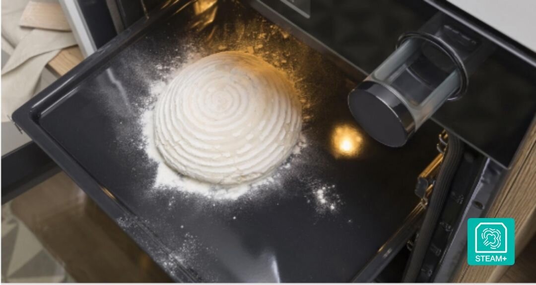 PIEKARNIK GORENJE BSA6737E15BG Surowy bochenek chleba uformowany w koszyku garowniczym leży na czarnej blasze posypanej mąką. Ikona Steam+ sugeruje użycie pary podczas wypieku dla lepszego wyrastania i chrupiącej skórki