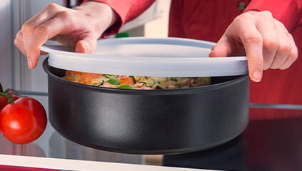Osoba zakłada białą pokrywkę na czarną patelnię z jedzeniem. Obok leży pomidor.