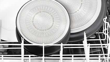 Dwie patelnie umieszczone w zmywarce, widoczne od spodu z metalicznymi, perforowanymi powierzchniami. Na powierzchni jednej z patelni widoczny jest tekst: \'TEFAL INDUCTION\'.