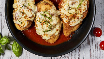 Na patelni znajdują się trzy kawałki zapiekanej potrawy z serem na sosie pomidorowym. Obok leżą liście bazylii i czerwone pomidorki koktajlowe.