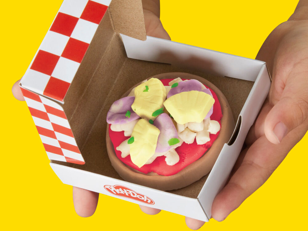 Na zdjęciu widać ręce trzymające małe pudełko z ciastolinową pizzą w środku. Pudełko ma wzór w czerwono-białą kratkę i logo \'Play-Doh\'.