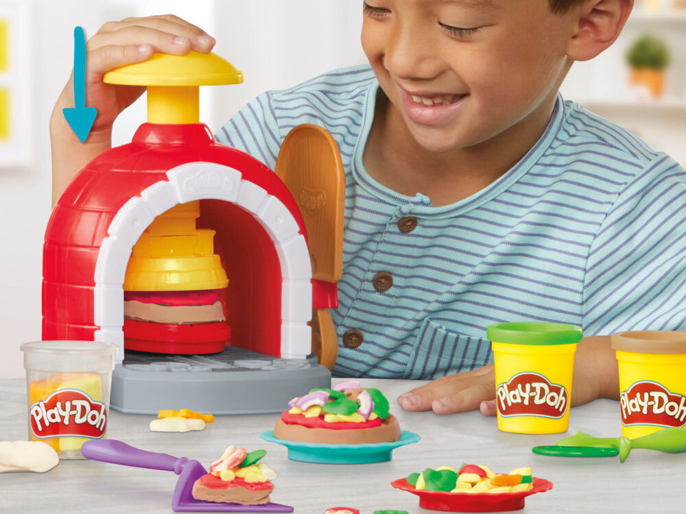 Dziecko bawi się zestawem do robienia pizzy z ciastoliny, w skład którego wchodzi piec, narzędzia i pojemniki z ciastoliną Play-Doh. Na stole widoczne są kolorowe elementy pizzy i akcesoria do zabawy.