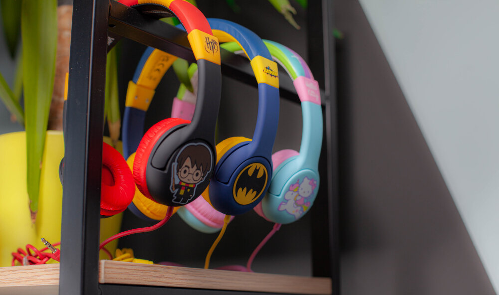 Na zdjęciu widoczne są trzy pary kolorowych słuchawek nausznych zawieszonych na metalowym stojaku. Słuchawki mają różne wzory, w tym motyw z postacią z okularami, logo nietoperza oraz jednorożca.
