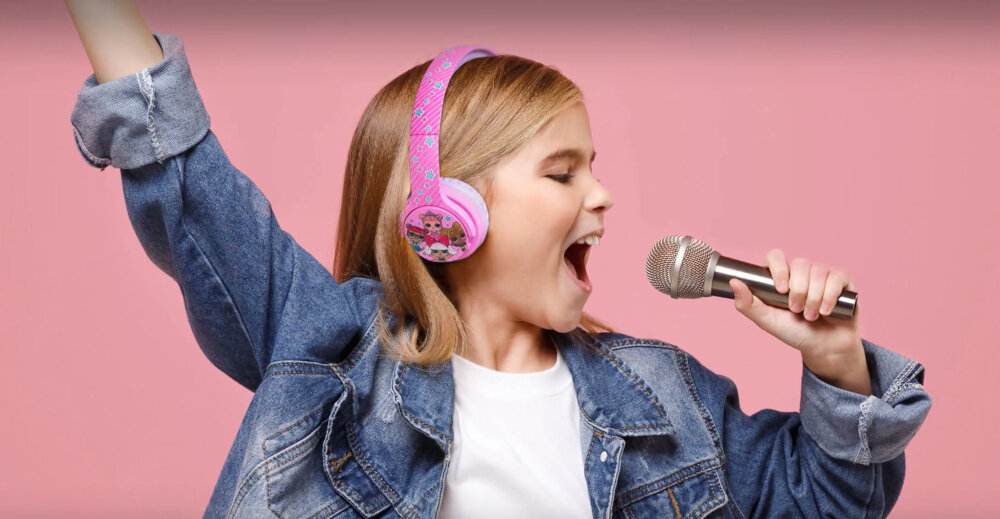 Dziecko w dżinsowej kurtce trzyma mikrofon i nosi różowe słuchawki z motywem bajkowym na różowym tle.