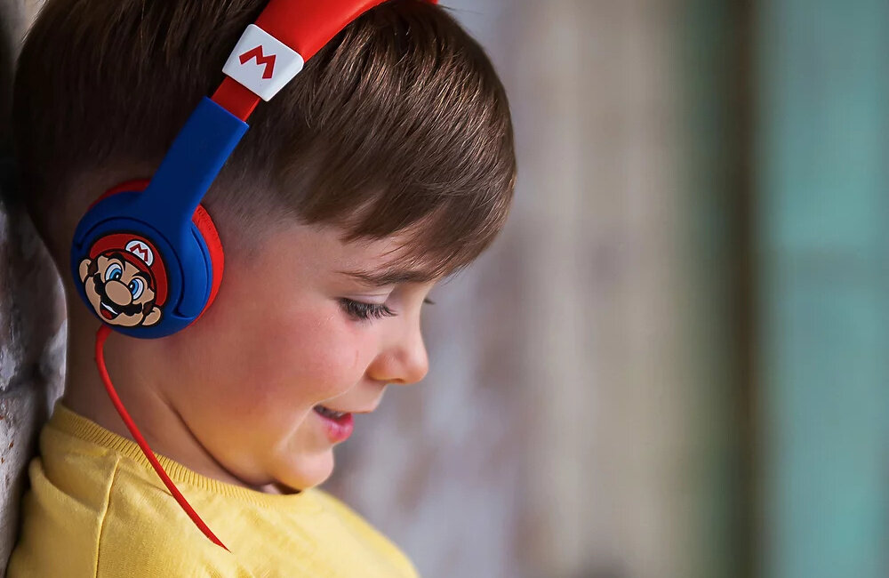 Dziecko nosi niebiesko-czerwone słuchawki nauszne z motywem Super Mario. Na pałąku widoczne jest logo z literą \'M\'.