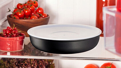 Czarna patelnia z białym wnętrzem znajduje się na półce w lodówce. W tle widoczne są miski z pomidorami i czerwonymi owocami.