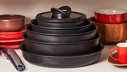 Na obrazie widoczny jest zestaw czarnych patelni ułożonych jedna na drugiej, obok czerwonych naczyń i filiżanki. Patelnie mają różne rozmiary, a na górze znajduje się pokrywka.