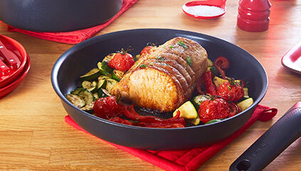 Na drewnianym blacie stoi czarna patelnia z uchwytem, w której znajduje się pieczone mięso z warzywami, takimi jak pomidory i cukinia. W tle widoczne są czerwone akcesoria kuchenne.