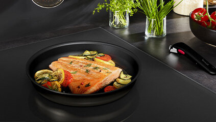 Na płycie kuchennej znajduje się patelnia z kawałkiem łososia i warzywami. Obok patelni leży odłączany uchwyt, a w tle widać zioła i miski z warzywami.