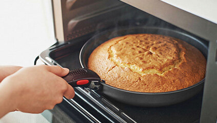 Osoba wyciąga z piekarnika ciasto w okrągłej patelni z odłączanym uchwytem. Patelnię trzymają ręce, a ciasto jest dobrze wypieczone i złociste.