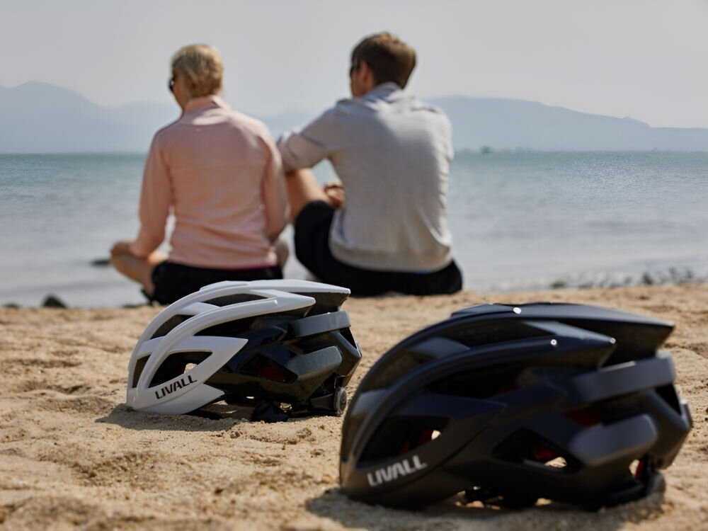 Kask rowerowy LIVALL BH60SE Neo Czarny Szosowy (rozmiar L) biały i czarny kask leżące na piasku w tle dwie osoby