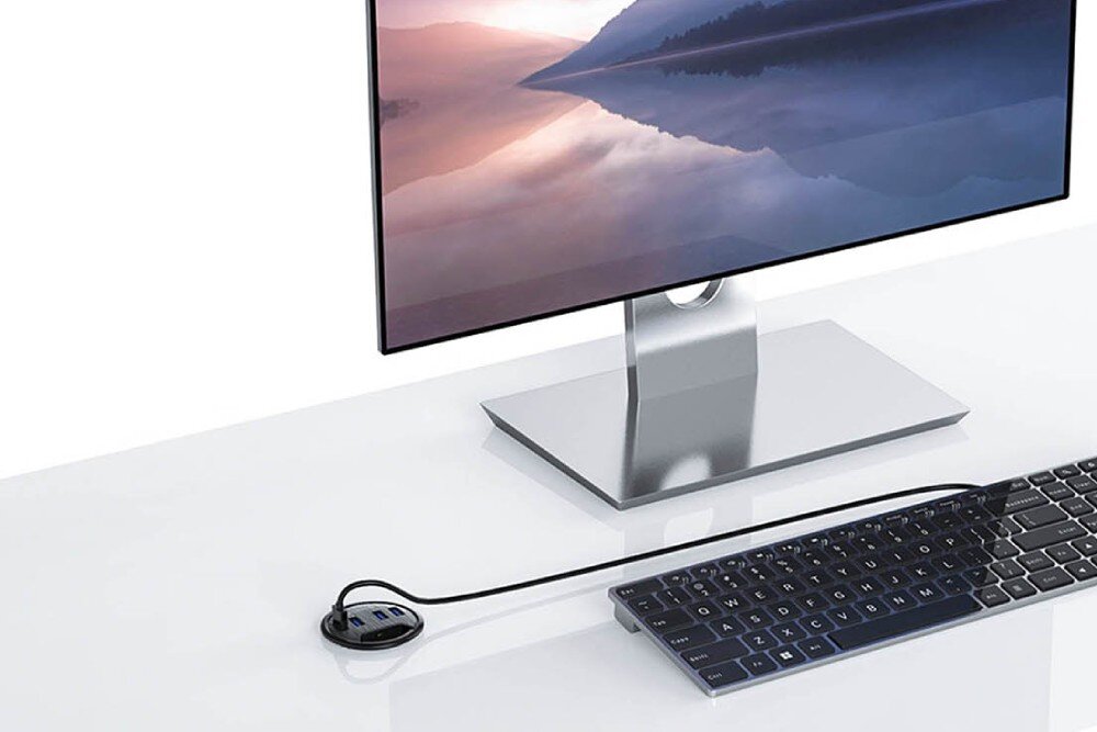 Hub ORICO DESK-4U-BK-BP Monitor z krajobrazem górskim na ekranie stoi na białym biurku obok cienkiej klawiatury, a obok znajduje się czarny okrągły hub USB z kilkoma portami, opis