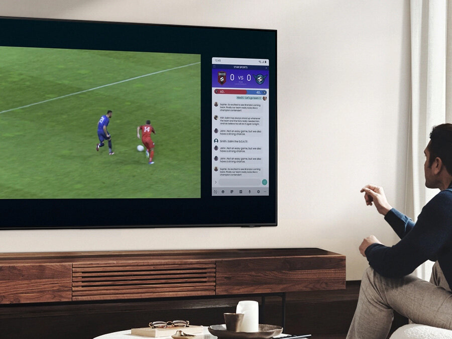 Мужчина использует QLED Multi View, чтобы одновременно смотреть футбольный матч и читать новости на одном экране.  Q77BATXXH